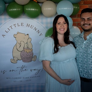May include: A backdrop featuring a light blue gingham pattern, a cartoon bear with a honey pot, and the text A LITTLE HUNNY is on the way! WELCOMING BABY FARRAH 03.28.2026, surrounded by a balloon arch in shades of green, blue, and cream.