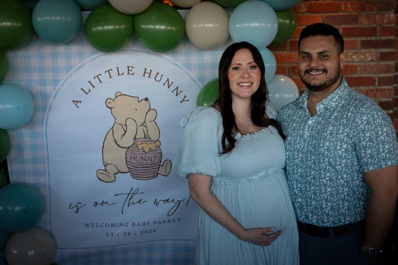 May include: A backdrop featuring a light blue gingham pattern, a cartoon bear with a honey pot, and the text A LITTLE HUNNY is on the way! WELCOMING BABY FARRAH 03.28.2026, surrounded by a balloon arch in shades of green, blue, and cream.