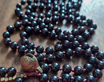 May include: A dark red gemstone mala necklace with a brass charm. The necklace is made of 108 beads and is strung on a red cord.