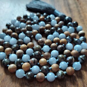 May include: A beaded mala necklace with a dark gray tassel. The necklace features a mix of light blue, dark brown, and wooden beads. The beads are strung together with a light blue cord. The necklace is displayed on a wooden surface.