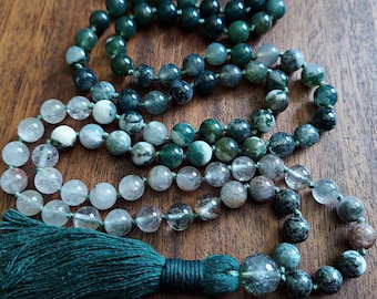 Può includere: Una collana mala di cristallo verde e trasparente con un fiocco verde. La collana è fatta di perline rotonde ed è infilata su un cordoncino verde.