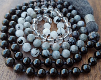 May include: A black and grey mala bead necklace with a black tassel. The beads are made of black onyx, clear quartz, and grey labradorite.