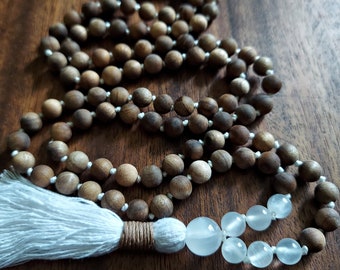 May include: A brown wooden mala bead necklace with a white tassel and clear quartz beads. The necklace is laid out on a wooden surface.
