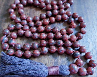May include: A mala necklace featuring reddish-brown, speckled beads. The beads are strung together with a matching cord and finished with a dark brown tassel. The beads are approximately 8mm in diameter.