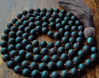 Può includere: Una collana mala in legno marrone scuro con un nappa. Le perle sono rotonde e lisce, e la nappa è fatta di tessuto marrone.