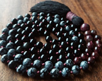 May include: A mala or prayer beads made of black obsidian, snowflake obsidian, and red jasper beads. The mala has a black tassel.