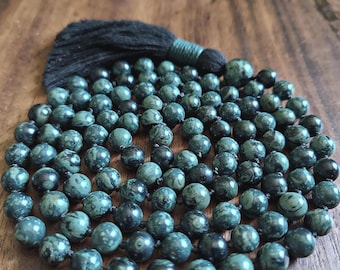 May include: A mala necklace featuring numerous round, dark green beads with black mottled patterns, strung together with visible knots between each bead. The necklace is finished with a single, thick black fabric tassel at the top.