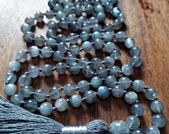 May include: A beaded necklace featuring round, gray-blue gemstones strung on a dark cord. The necklace has a gray tassel and a silver-colored metal accent. The beads are approximately 0.3 inches in diameter.