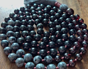 May include: A black and grey beaded mala necklace with a dark grey tassel. The beads are strung on a dark red cord.