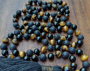 May include: A beaded necklace featuring alternating round tiger eye beads, smooth black obsidian beads, and porous black lava stone beads. The necklace is finished with a long, thick black thread tassel secured by a wrapped black thread knot.