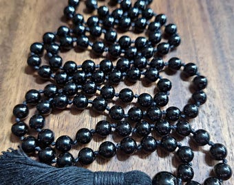 May include: A long necklace featuring numerous smooth, round, glossy black beads strung together with visible knots between each bead. The necklace terminates in a single, larger black bead attached to a thick, textured black fabric tassel.