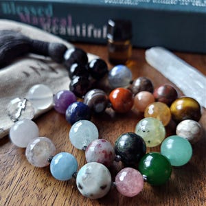 May include: A collection of colorful gemstone beads in various sizes and colors, including pink, green, blue, and orange. The beads are arranged in a circular pattern on a wooden surface. A book with the words "Blessed Magical Beautiful" is in the background.