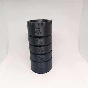 May include: A black, cylindrical stacking toy with the numbers 2 through 5 printed on the sides. The top piece has a ridged edge and a slot. The toy is made of a matte black material and sits on a white surface.