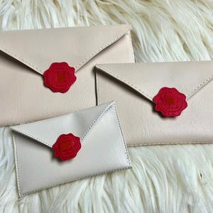 May include: Three beige envelope-shaped pouches with red wax seals. The pouches are arranged in a pyramid shape, with the largest pouch at the bottom and the smallest pouch at the top.