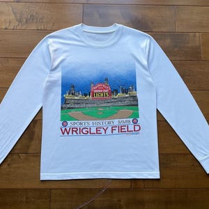 Pode incluir: Camiseta branca de manga comprida com um gráfico colorido do estádio de beisebol Wrigley Field. O gráfico inclui o texto "SPORTS HISTORY 8/8/65" e "WRIGLEY FIELD". A camiseta tem gola redonda e mangas compridas.