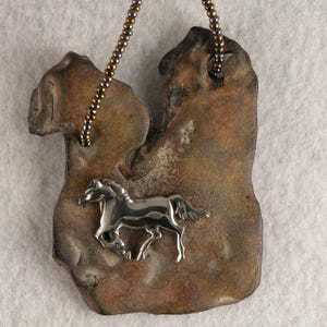 Può includere: Un ciondolo rustico con un ciondolo a forma di cavallo argentato su una base marrone di forma organica. Il ciondolo è sospeso da un cordoncino di perline. Il ciondolo a forma di cavallo è dettagliato e sembra essere in metallo lucido.