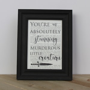 Puede incluir: Una impresión enmarcada con fondo blanco y negro que presenta una cita de un libro. La cita dice "You're an absolutely stunning murderous little creature." La cita está resaltada en negro y un pequeño gráfico de cuchillo se encuentra debajo del texto.