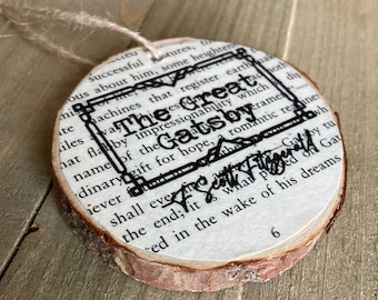 Può includere: Un ornamento rotondo in legno con sfondo bianco e testo nero. Il testo recita "The Great Gatsby" con un bordo decorativo intorno. L'ornamento è decorato con uno spago di iuta marrone.
