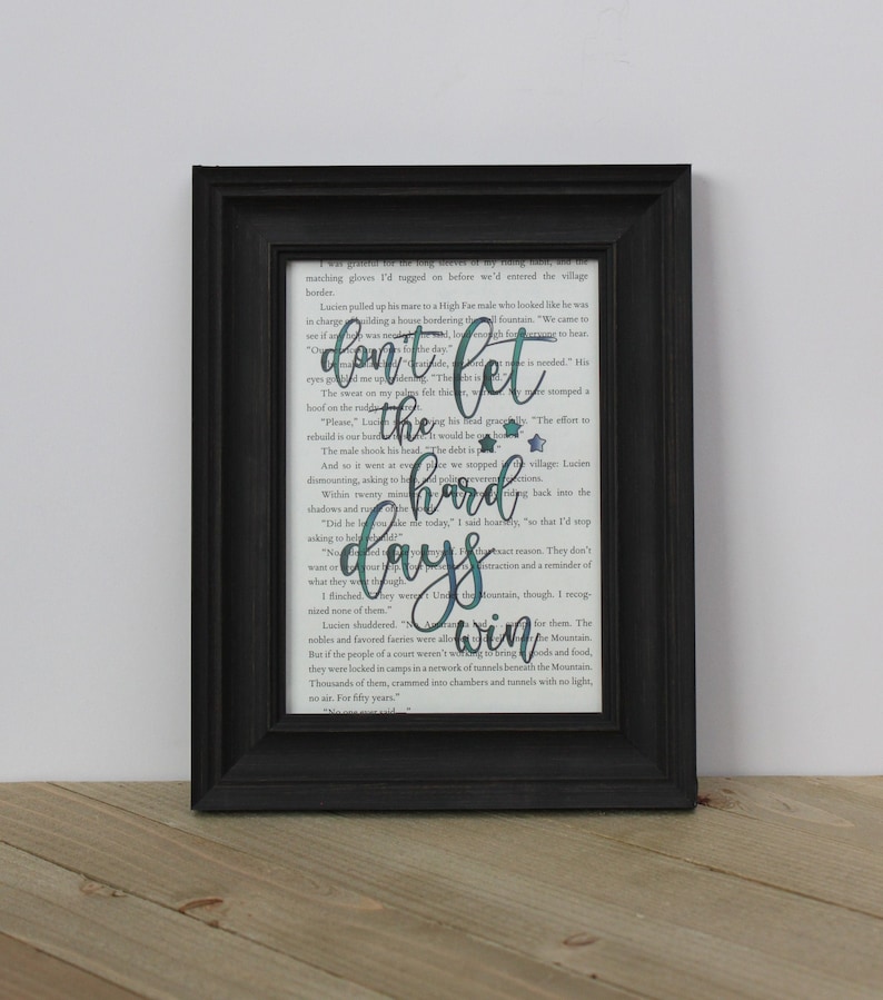 May include: A framed print with a black frame featuring the quote "Don't let the hard days win" in teal script over a book page background. The print includes small teal stars.