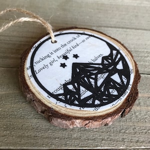May include: A wooden round ornament with a black and white design of a crescent moon and stars. The ornament is decorated with text from a book, including the phrase "Lovely girl, beautiful bird - so sweet".