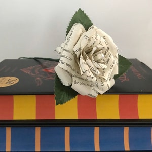 May include: A handmade paper rose made from book pages rests atop a stack of colorful books. The rose is white with visible text, and green leaves. The books have painted stripes in red, yellow, blue, and green.