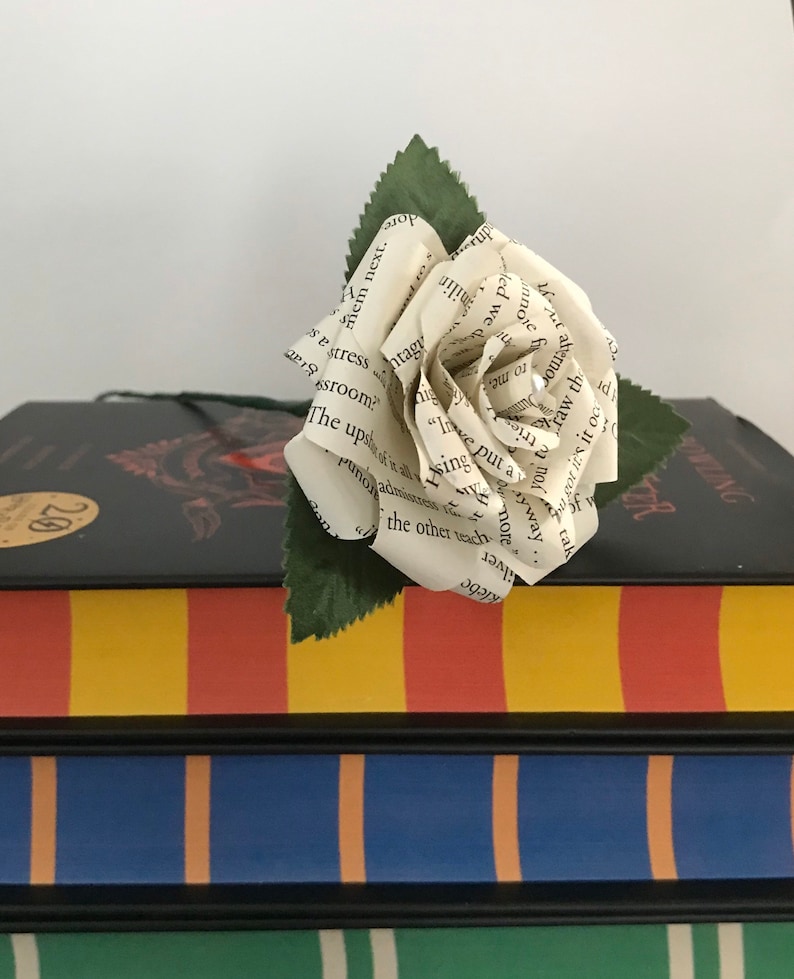 May include: A handmade paper rose made from book pages rests atop a stack of colorful books. The rose is white with visible text, and green leaves. The books have painted stripes in red, yellow, blue, and green.