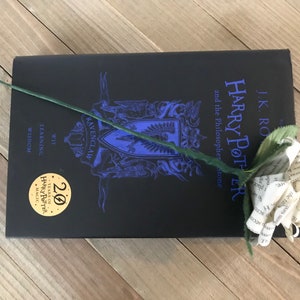 May include: A black hardcover book titled "Harry Potter and the Philosopher's Stone" with a paper flower on top. The book has a blue Ravenclaw crest and a gold 20 Years of Harry Potter Magic sticker.