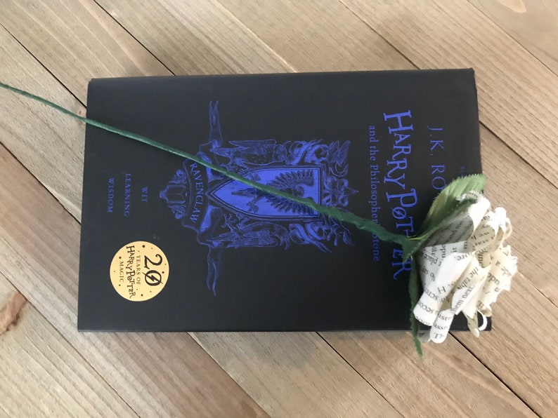 May include: A black hardcover book titled "Harry Potter and the Philosopher's Stone" with a paper flower on top. The book has a blue Ravenclaw crest and a gold 20 Years of Harry Potter Magic sticker.