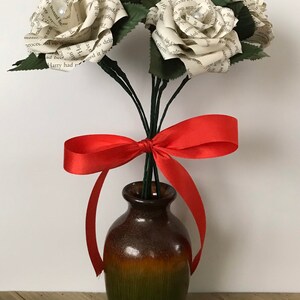 May include: A bouquet of paper roses made from book pages, with green leaves and stems, tied with a red satin ribbon. The flowers are in a brown and green ceramic vase, set against a light background.