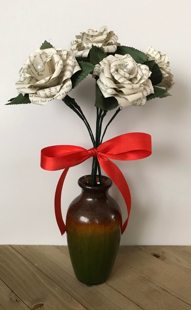 May include: A bouquet of paper roses made from book pages, with green leaves and stems, tied with a red satin ribbon. The flowers are in a brown and green ceramic vase, set against a light background.