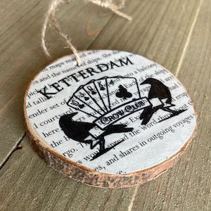 May include: A round wooden ornament with a printed image of a crow holding playing cards and a book with the text "Crow & Quill" and "Ketterdam".