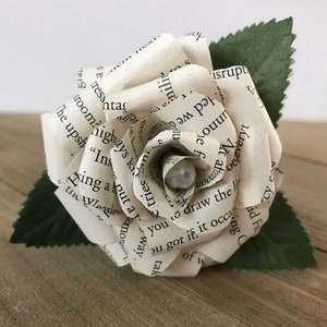 May include: A handmade paper rose with pages from a book, featuring a pearl center and green leaves. The white petals display printed text, creating a unique floral design. This craft item is suitable for home decor or as a gift.