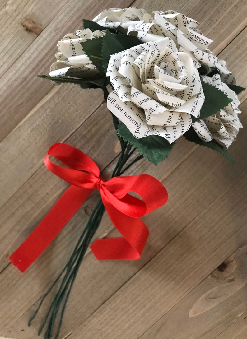 May include: A bouquet of paper roses made from book pages, with green leaves and stems. The bouquet is tied with a bright red ribbon, creating a unique and literary floral arrangement.