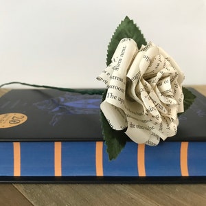May include: A handmade paper rose with a green stem and leaves rests on a closed book. The rose is crafted from pages of a book, displaying visible text. The book has a blue cover with orange stripes and a black cover with a gold sticker.