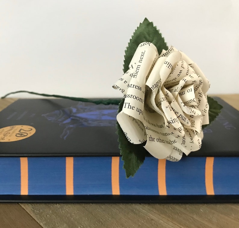 May include: A handmade paper rose with a green stem and leaves rests on a closed book. The rose is crafted from pages of a book, displaying visible text. The book has a blue cover with orange stripes and a black cover with a gold sticker.