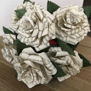 May include: A bouquet of paper roses made from book pages. The roses are white with printed text, and the green leaves add a natural touch. Each rose has a pearl accent. The bouquet is arranged on a wooden surface.