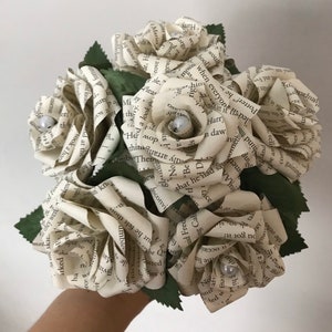 May include: A bouquet of paper roses made from book pages. Each cream-colored rose has a pearl center and is surrounded by green paper leaves. The text from the book pages is visible on the petals.