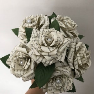 May include: A bouquet of paper roses, crafted from book pages, with pearl accents in the center of each bloom. Green paper leaves are interspersed among the flowers. The bouquet is held against a white background.