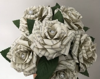 Puede incluir: Un ramo de rosas de papel, hechas con páginas de libros, con acentos de perlas en el centro de cada flor. Hojas de papel verde oscuro se intercalan entre las flores. El ramo se sostiene sobre un fondo blanco.