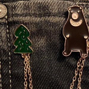 Könnte beinhalten: Zwei Emaille-Anstecknadeln: ein grüner Weihnachtsbaum und ein schwarzer Bär mit einem lächelnden Gesicht. Beide Anstecknadeln haben goldfarbene Ketten. Die Anstecknadeln werden auf einem Jeanshintergrund präsentiert.