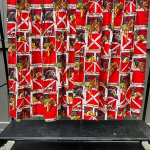 May include: Red fabric curtain with a repeating pattern of horses in stalls. The stalls are red with white cross-buck designs. The horses are various shades of brown, black, and gray. The curtain is hanging from a metal rod with white clothespins.