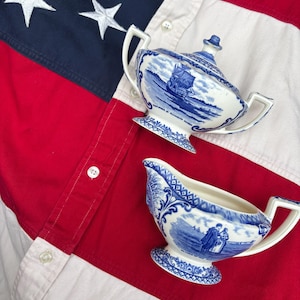 May include: A white ceramic sugar bowl with a lid and a matching creamer, both featuring blue transferware illustrations of a ship and figures. The sugar bowl is labeled Mayflower in Plymouth Harbor. The items are placed on a red, white, and blue fabric.