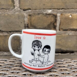 May include: White ceramic mug with a red stripe at the top and bottom. The mug features a black and white cartoon illustration of two figures and the text "love is... putting her before everything."