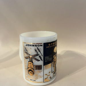May include: White ceramic mug with gold and black graphics. The mug features the text "JOHNSON" and "SKY LAB" along with an illustration of a space station. The other side has the text "SPACE HOUSTON, TEXAS" and an astronaut on the moon. A vintage souvenir.