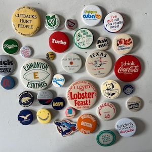 Puede incluir: Una colección de botones vintage con varios diseños y lemas. Algunos de los botones presentan logotipos, como los Edmonton Eskimos, Coca-Cola y Air Canada. Otros tienen dichos humorísticos, como "Tengo todo lo que realmente necesitas" y "Presiona este botón y mis pantalones se caerán."