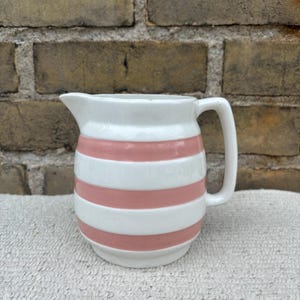 Peut inclure: Pichet en céramique blanche avec une anse et un bec verseur, orné de rayures horizontales roses. Le pichet est posé sur un fond texturé, présentant un design classique, idéal pour servir des boissons. Hauteur environ 15 cm.