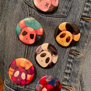 Könnte beinhalten: Sechs runde Anstecknadeln mit Comic-Tiergesichtern. Die Knöpfe befinden sich auf einer blauen Jeansjacke. Die Tiere sind ein rosa Hund mit schwarzen Augen, ein gelber Hund mit schwarzen Augen, ein weißer Hund mit schwarzen Augen, ein rosa Hund mit lila Augen, ein roter Hund mit schwarzen Augen und ein rosa Hund mit schwarzen Augen und X für Augen.