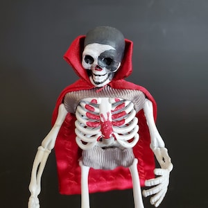 May include: A skeleton figure wearing a red cape with a painted skull head. The skull has black and white paint and a red mouth. The skeleton has a red painted heart on its chest.