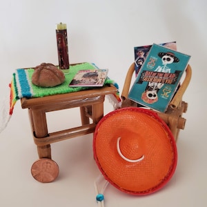May include: A miniature Day of the Dead scene with a small table, a chair, a sombrero, a candle, a sweet bread, and a Day of the Dead poster. The table has a green and blue striped tablecloth and a small magazine on top. The chair is made of bamboo and has a Day of the Dead poster on the back. The sombrero is orange and has a white band around it. The candle is yellow and has a religious image on it. The sweet bread is round and brown. The poster is blue and has a skeleton on it. The poster says "Day of the Dead" and "Dia de Muertos".