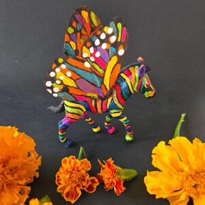 May include: A colorful, hand-painted figurine of a zebra with butterfly wings. The zebra has rainbow stripes, and the wings are a vibrant mix of orange, yellow, blue, and white. Marigold flowers are scattered around the figurine.